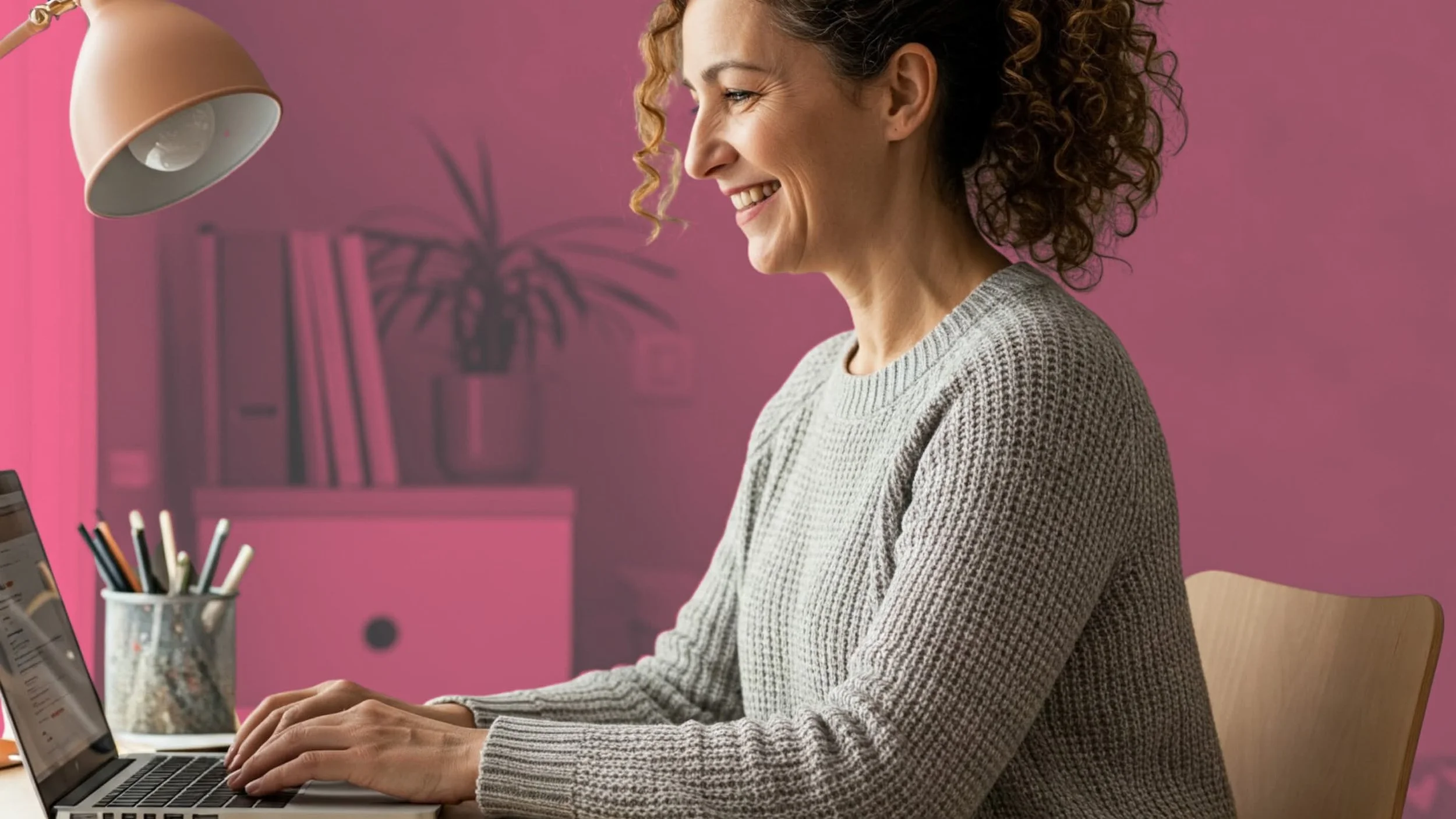 A woman with curly hair sitting at a desk, smiling, working on a laptop with a pink wall in the background.