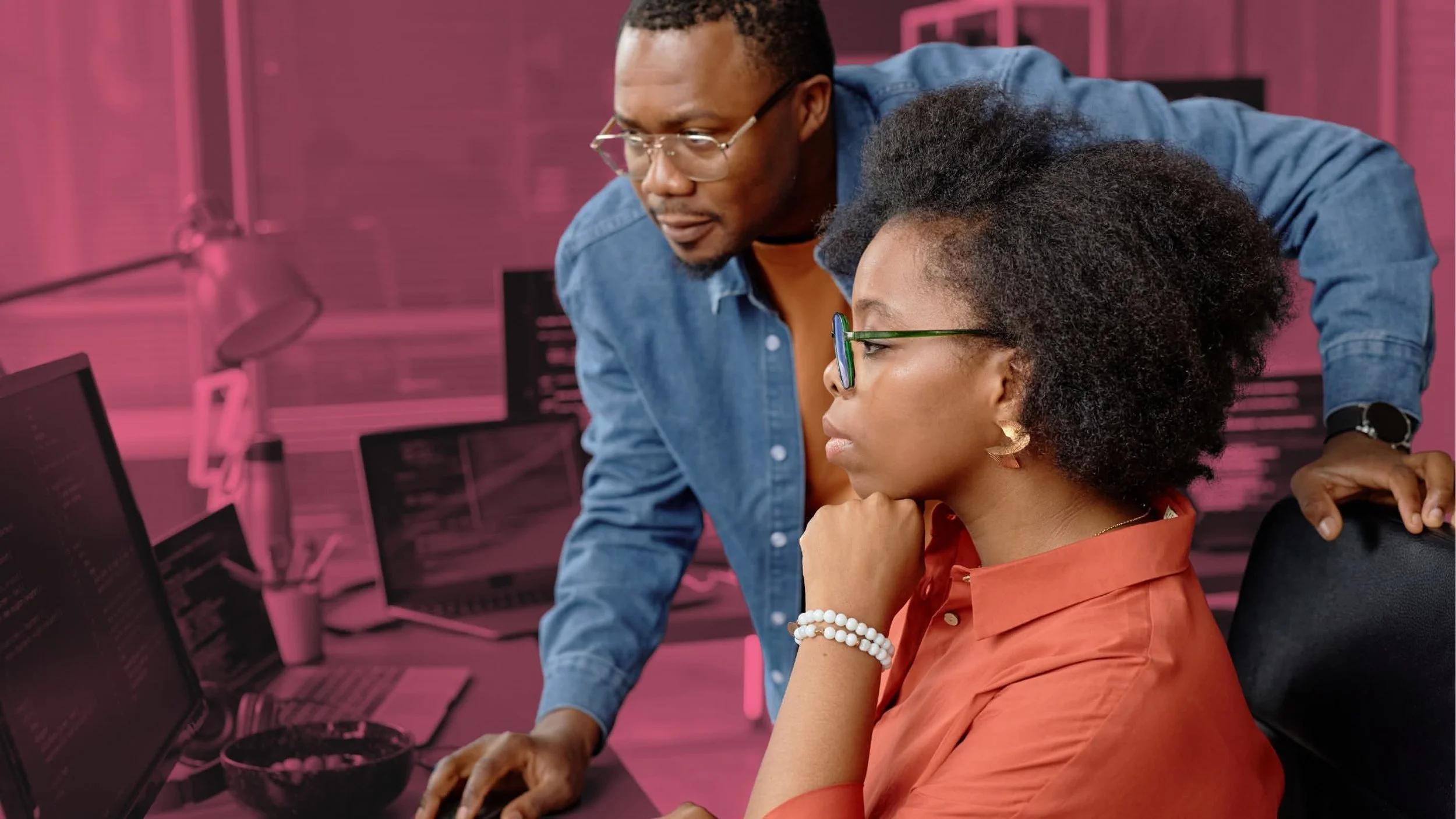 A man wearing glasses and a denim shirt is standing next to a woman with glasses, a orange shirt, and earrings, working on multiple computer monitors in a pink-toned office. The woman is focused on her screen, while the man is looking at her computer.