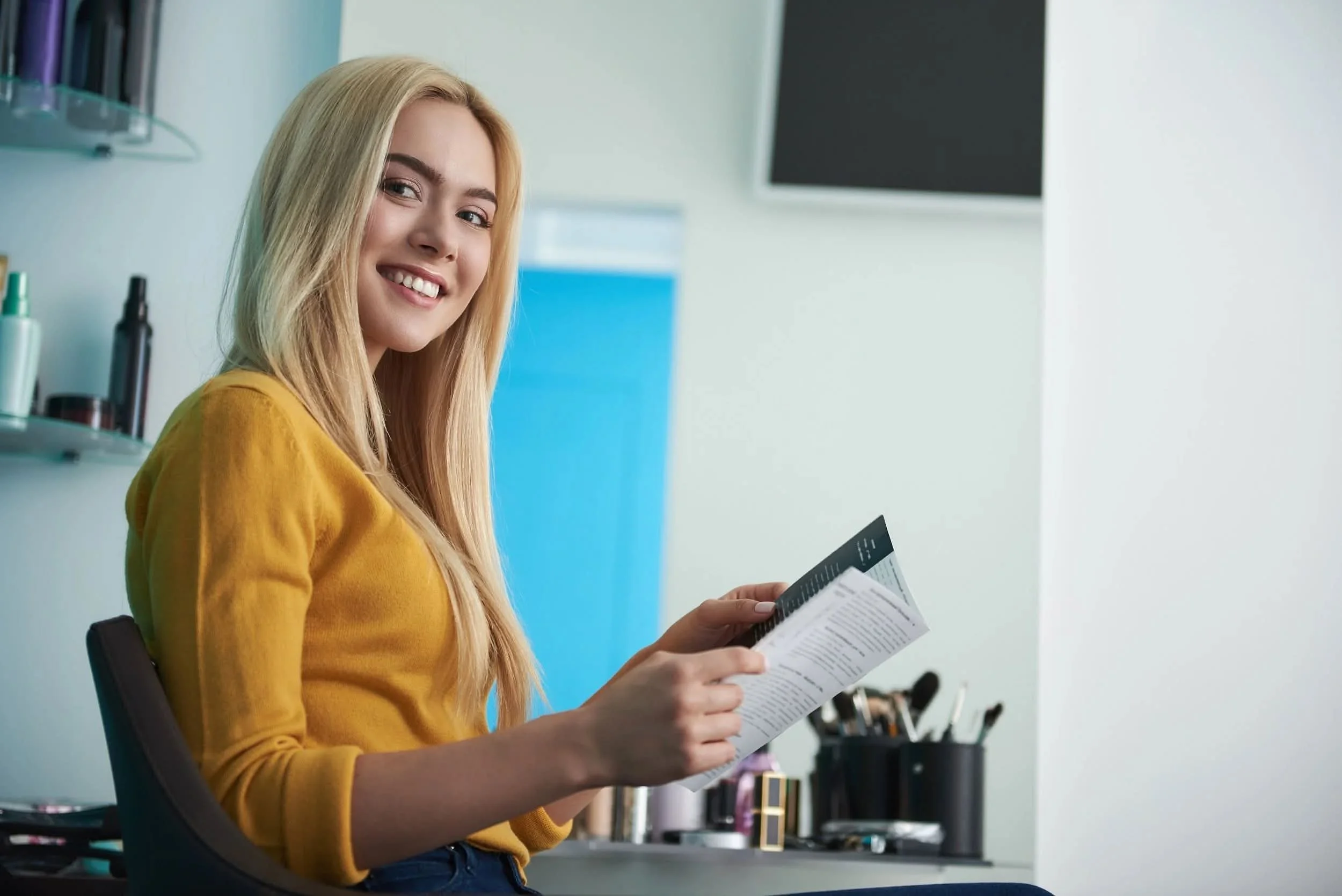A young woman with long blonde hair, wearing a yellow top, sitting in a salon or spa, smiling and holding a brochure or magazine, with hair products and brushes on a counter behind her.