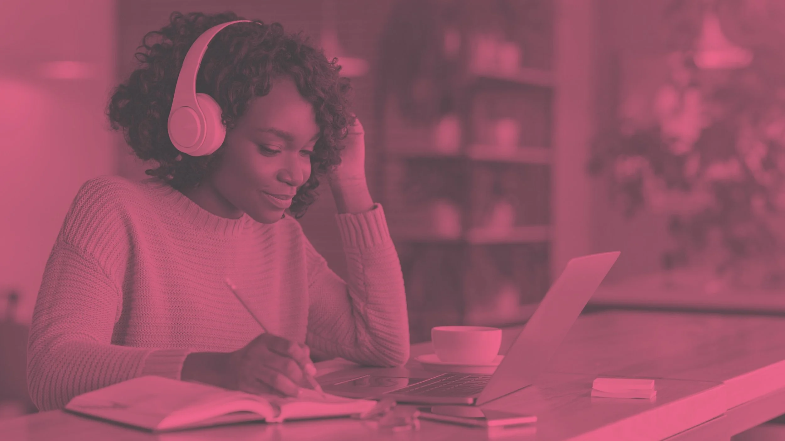 A woman with curly hair wearing pink headphones, sitting at a desk with a laptop, notebooks, and a cup, working or studying in a cozy indoor environment.