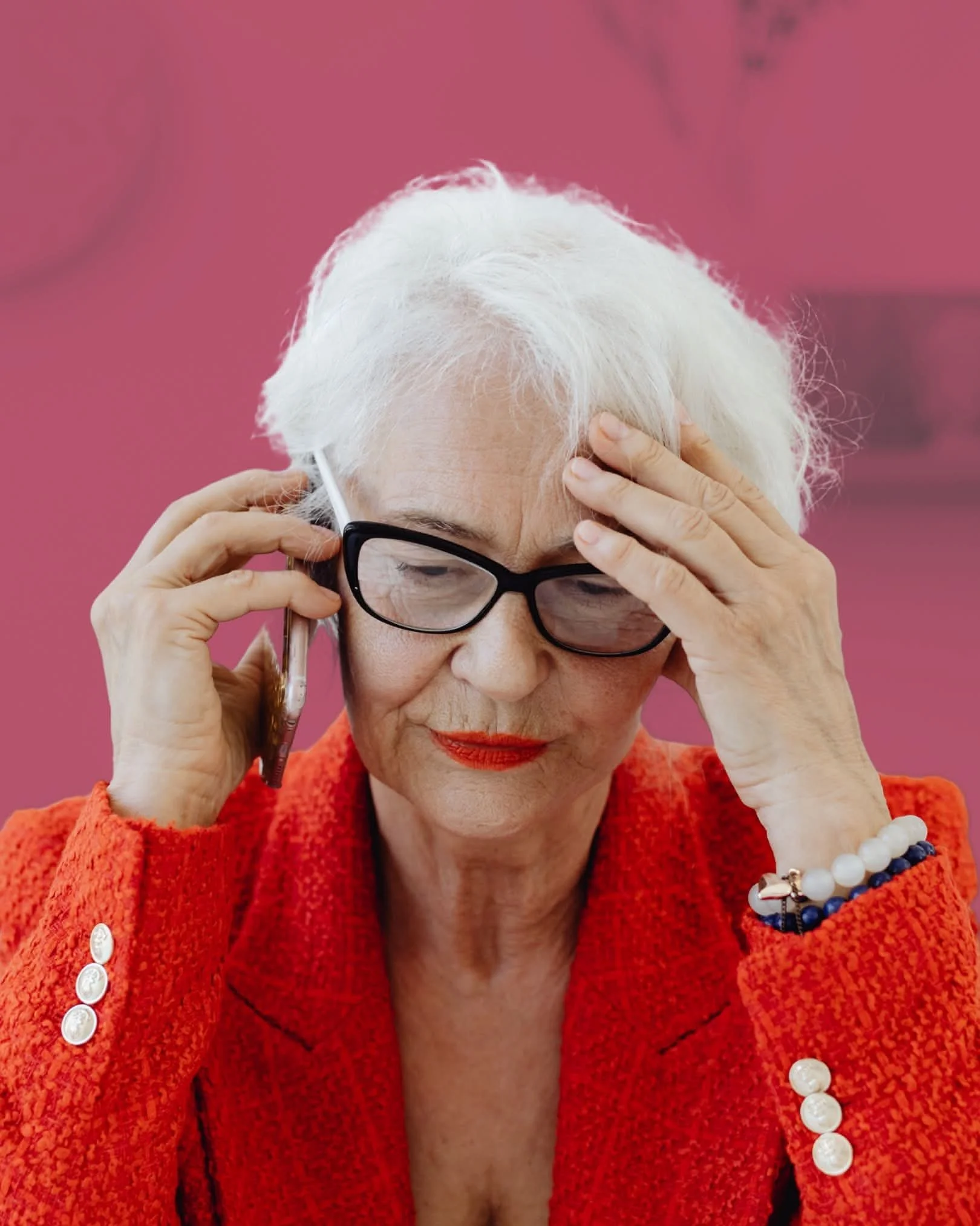 An elderly woman with white hair, wearing glasses and a red textured jacket, talking on the phone, with a pink background.