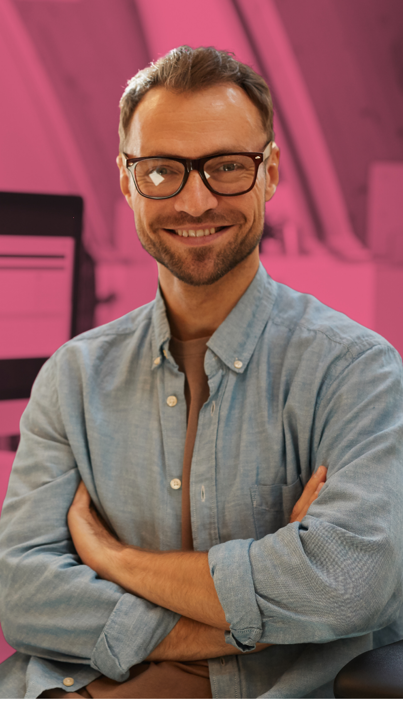 A smiling man with glasses and a beard, wearing a light blue button-up shirt, sitting with arms crossed in an indoor space with a pink background and blurred computer screens.