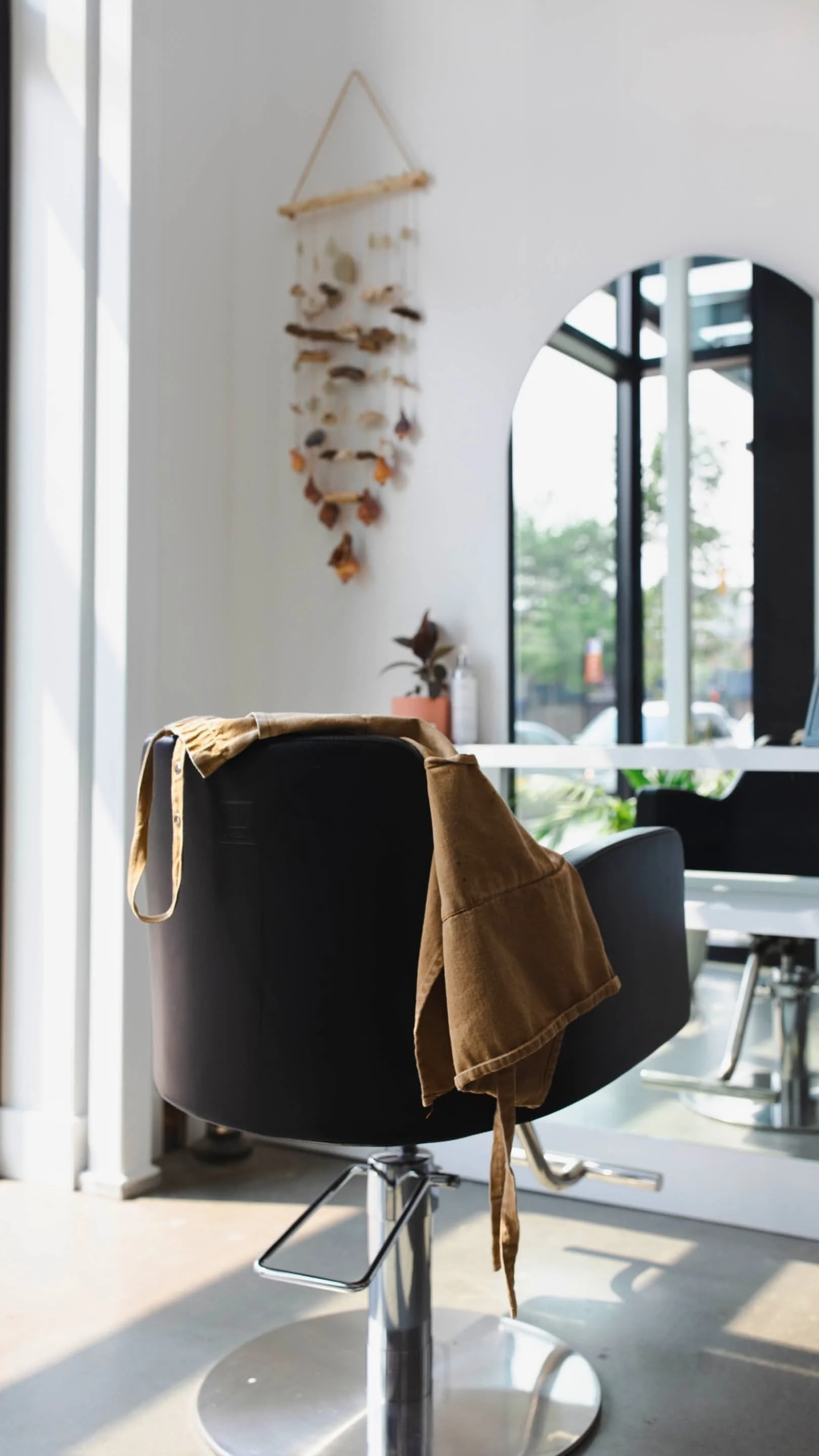 Interior of a modern hair salon with a black salon chair draped with a brown cape, a small potted plant on the table, a wall hanging, and large windows letting in natural light.