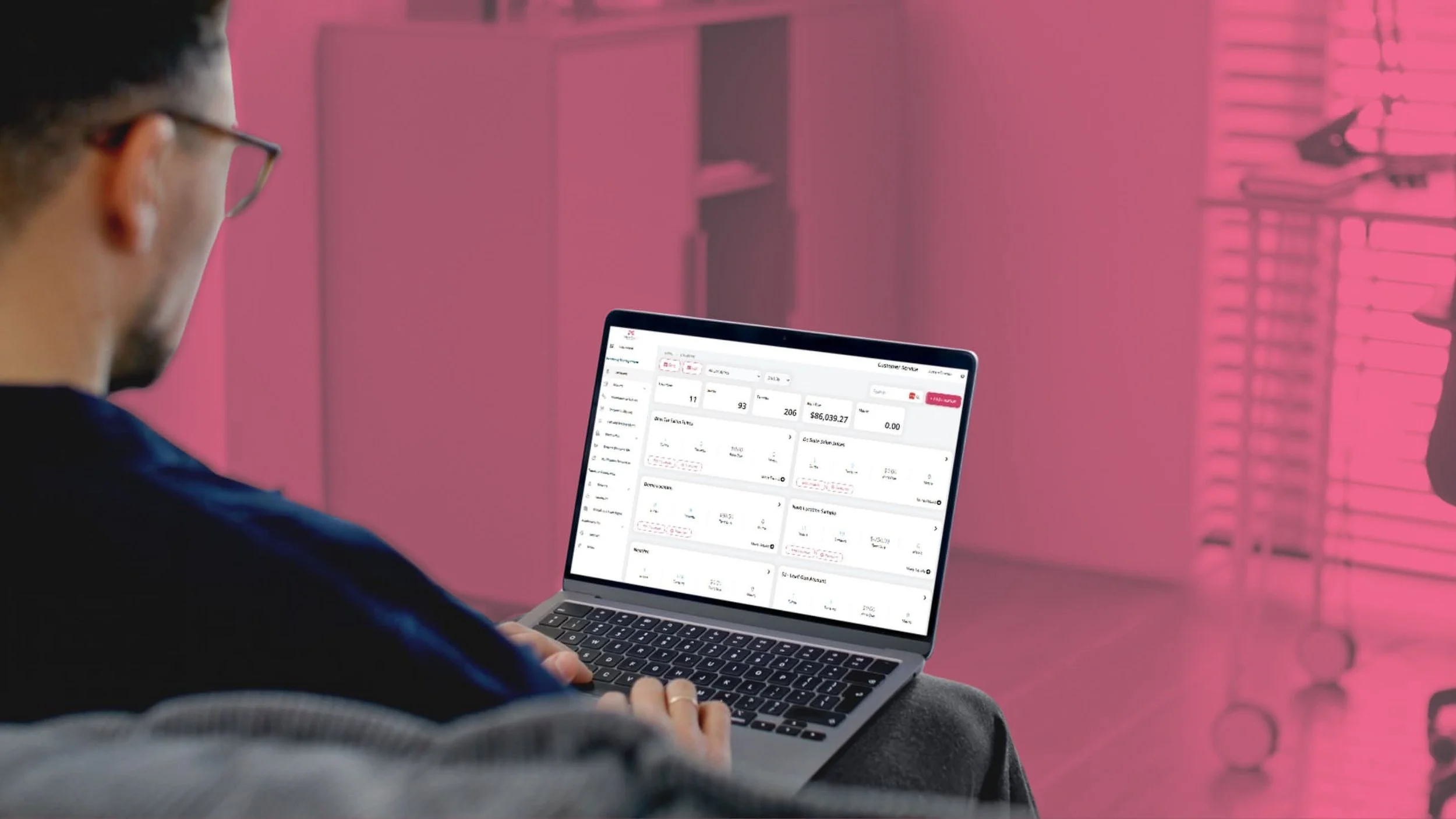 Person with glasses using a laptop with financial software on the screen, in a pink-lit room with shelves and a rolling cart in the background.