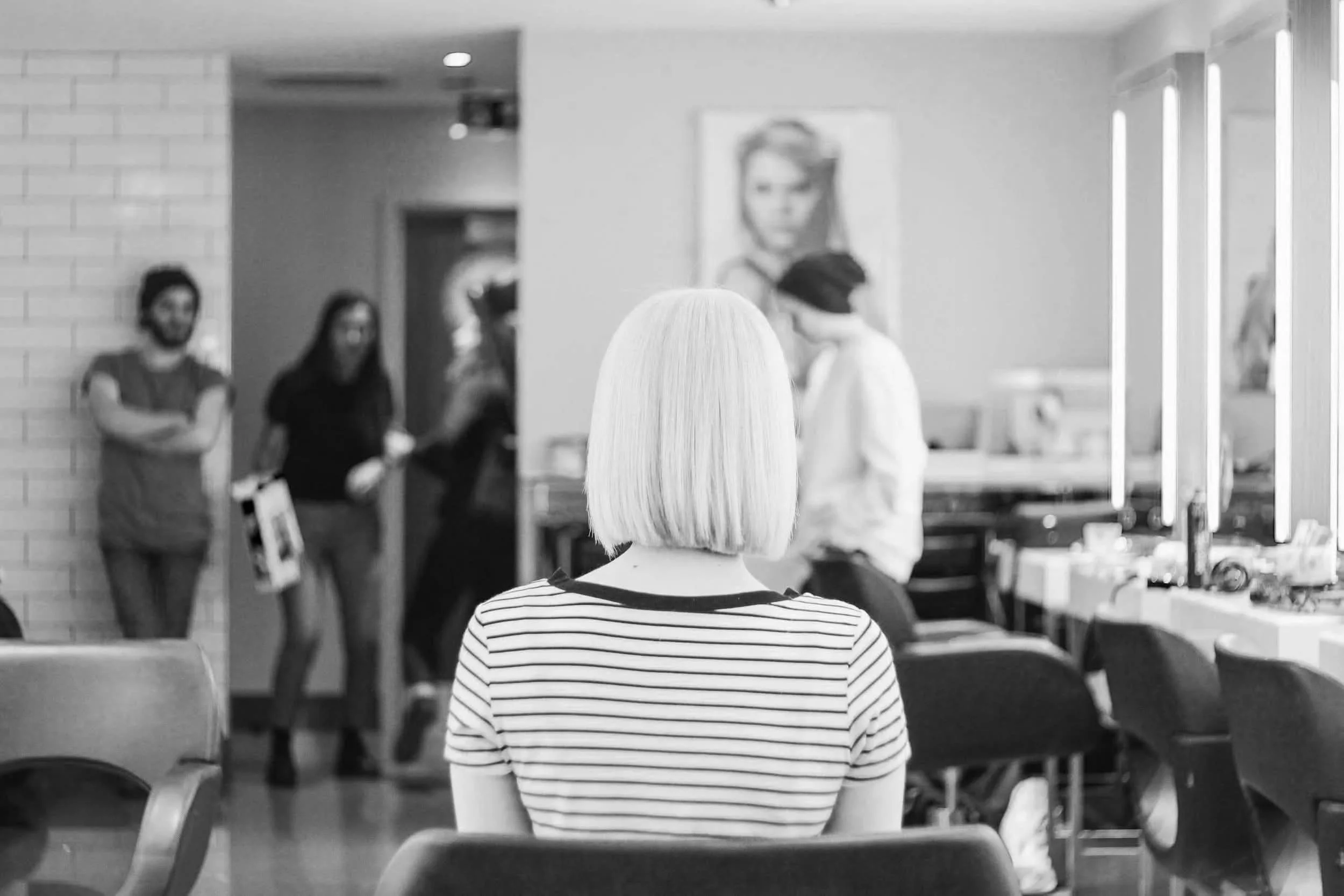 A woman with straight, shoulder-length blonde hair sitting in a room, facing away from the camera, with a group of people and a portrait on the wall in the background.