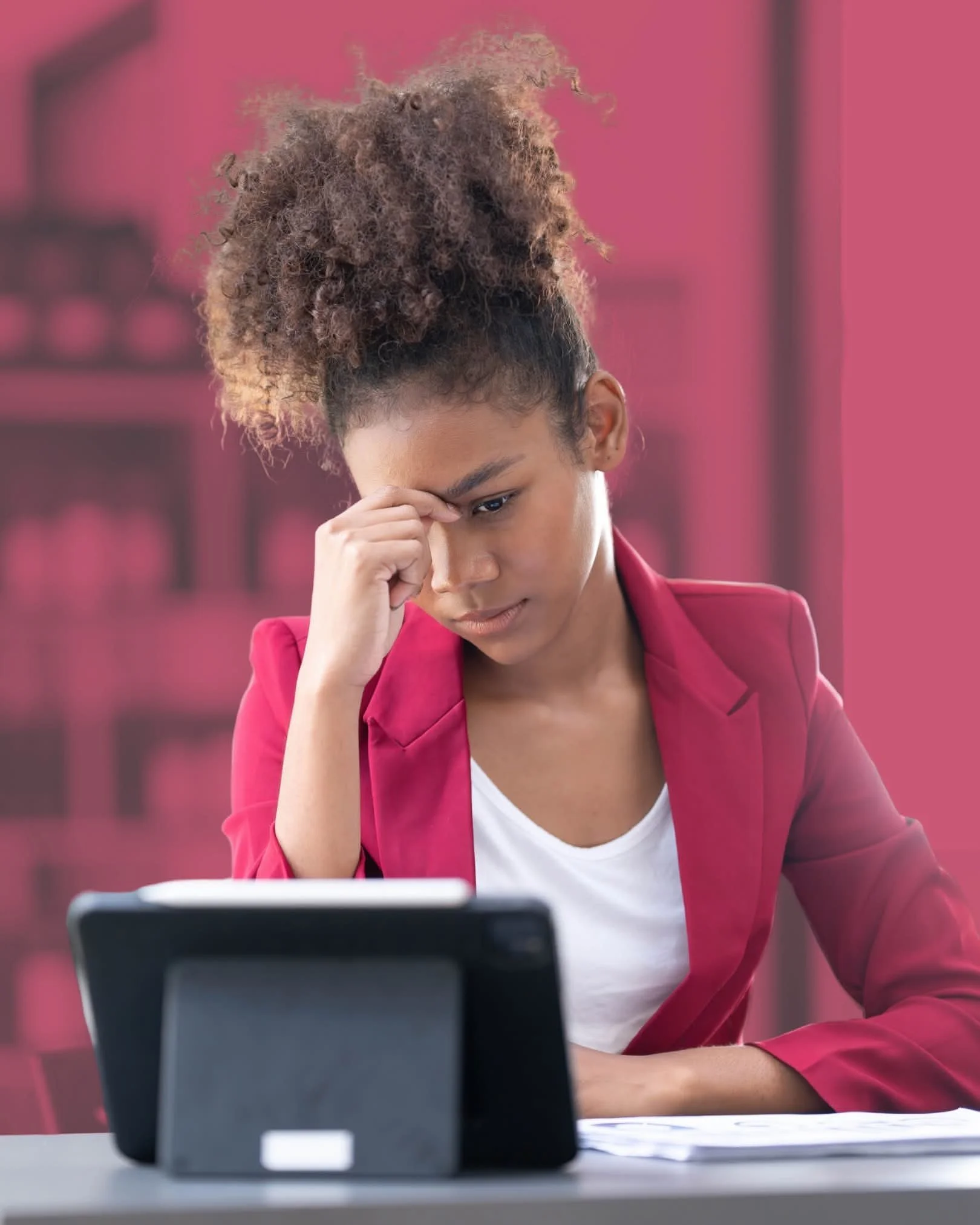 A young woman with curly hair in a high ponytail, wearing a pink blazer and white shirt, sitting at a desk and looking at a tablet device with a worried expression.