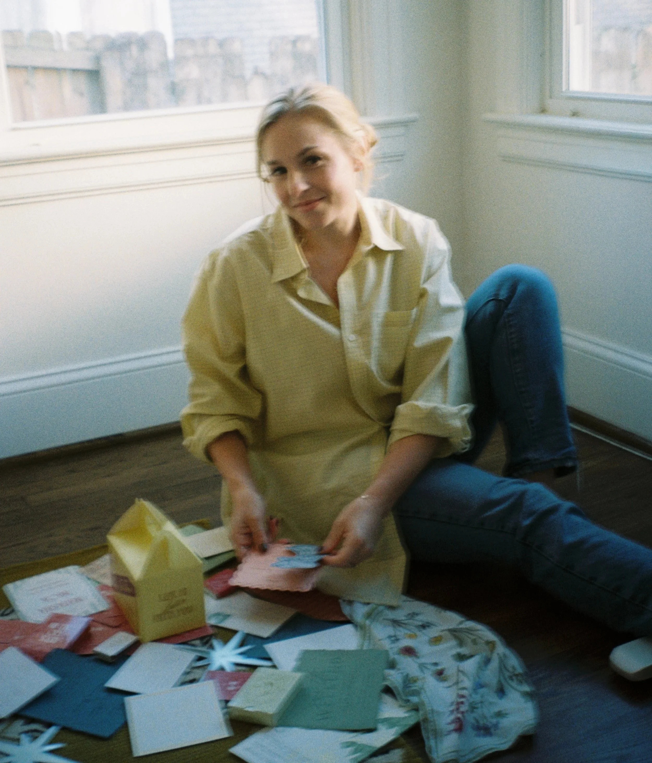 A woman sitting on the floor near a window, smiling, surrounded by colorful paper crafts and greeting cards.