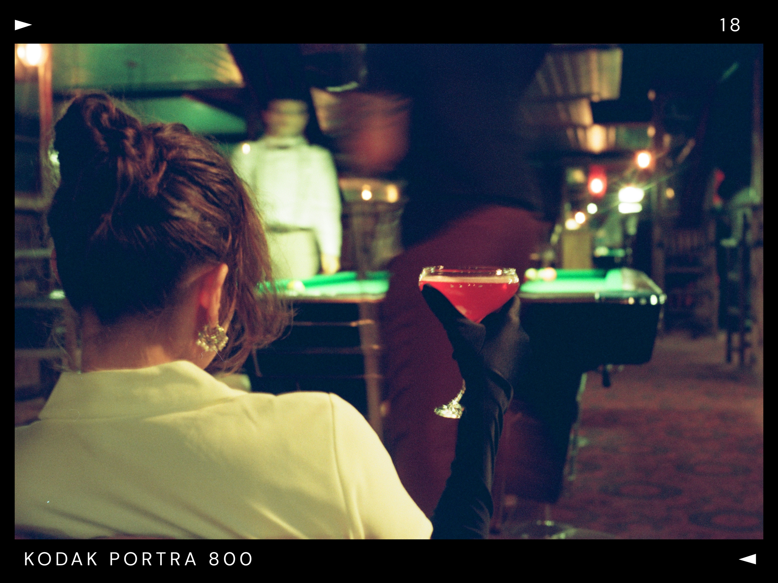 A woman with brown hair tied in a bun, wearing a yellow top and large earrings, holding a cocktail glass with a red drink in a dimly lit bar or pool hall with a green felt pool table and a man in a dark shirt and red pants walking by.