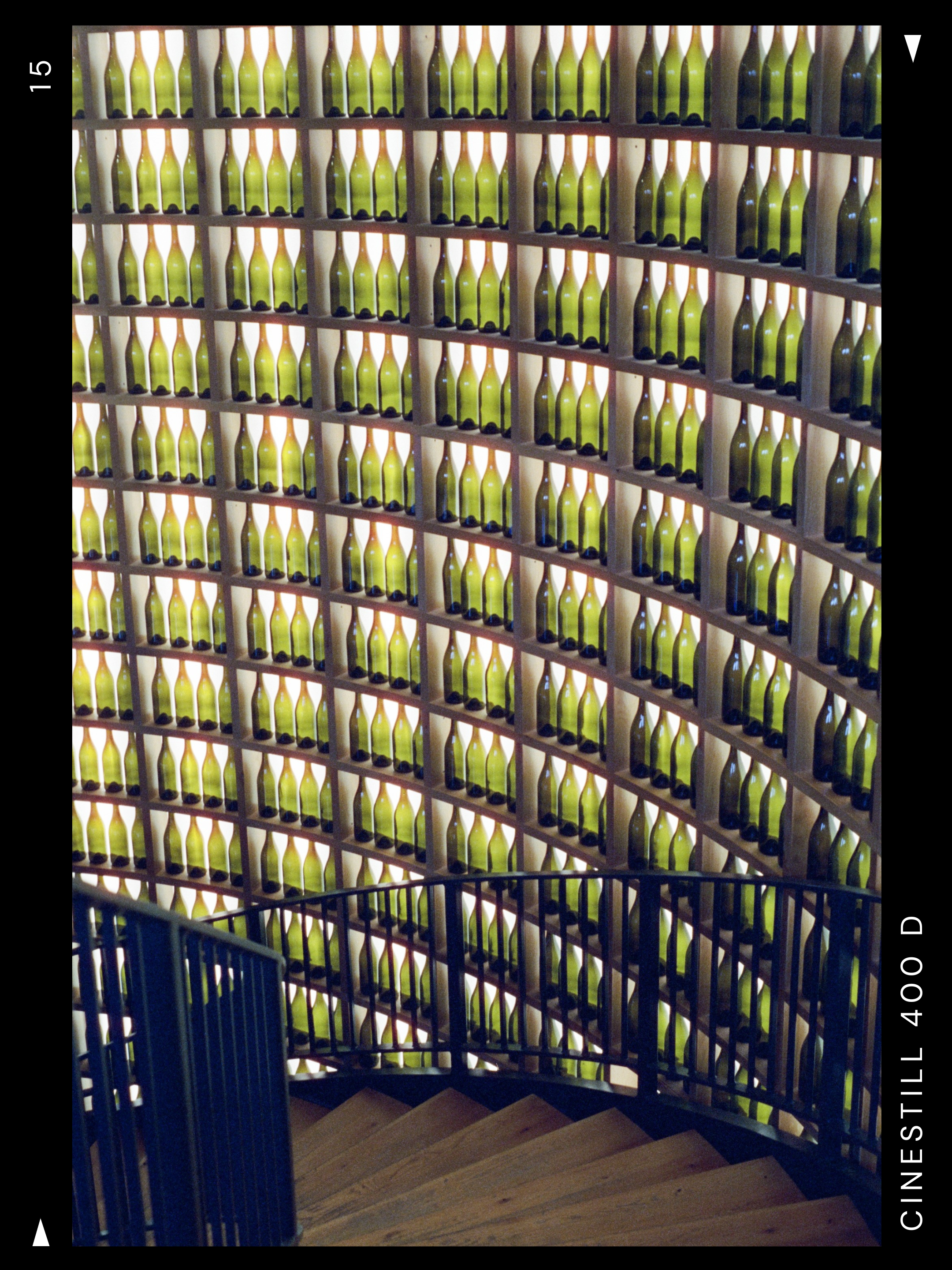 An interior shot of a spiral staircase with a black handrail and wooden steps, overlooking a wall made of numerous wine bottles arranged in rows within a wooden frame.