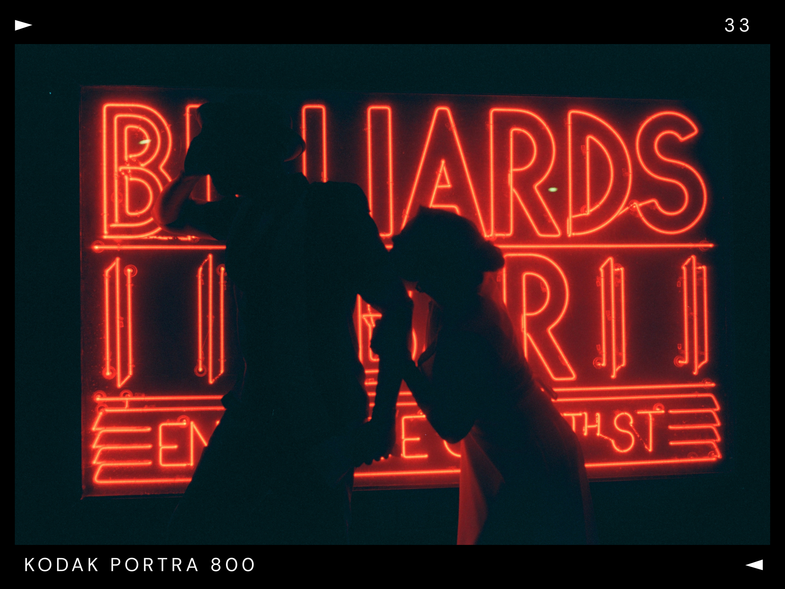 Silhouettes of a man and a woman kissing in front of a bright red neon sign that reads 'BILLIARDS' and 'EST.'