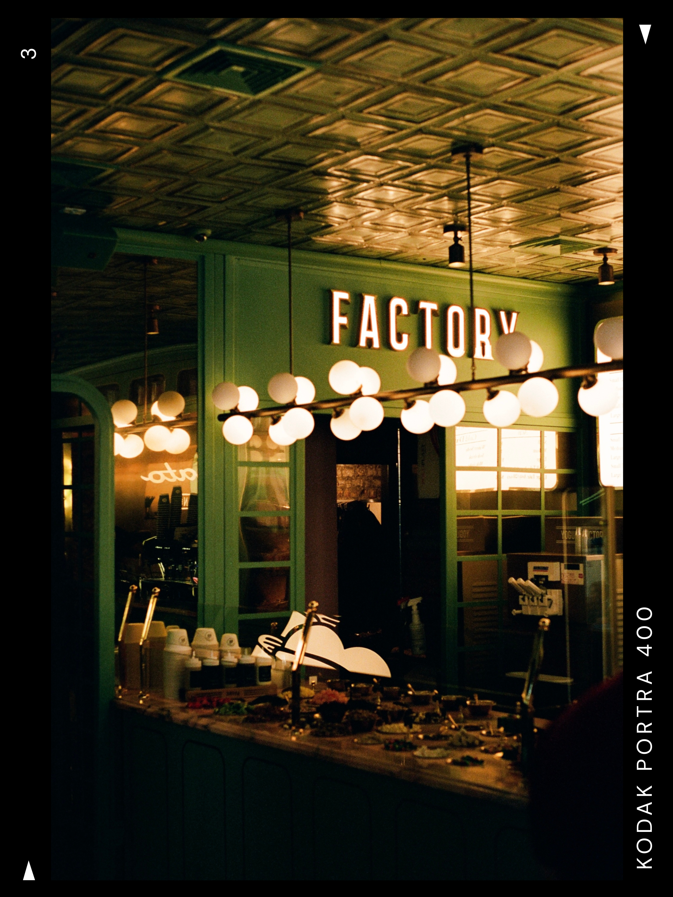 Indoor scene with a green wall displaying a bright 'Factory' sign, illuminated spherical pendant lights hanging from the ceiling, and a table with food or samples and cups.
