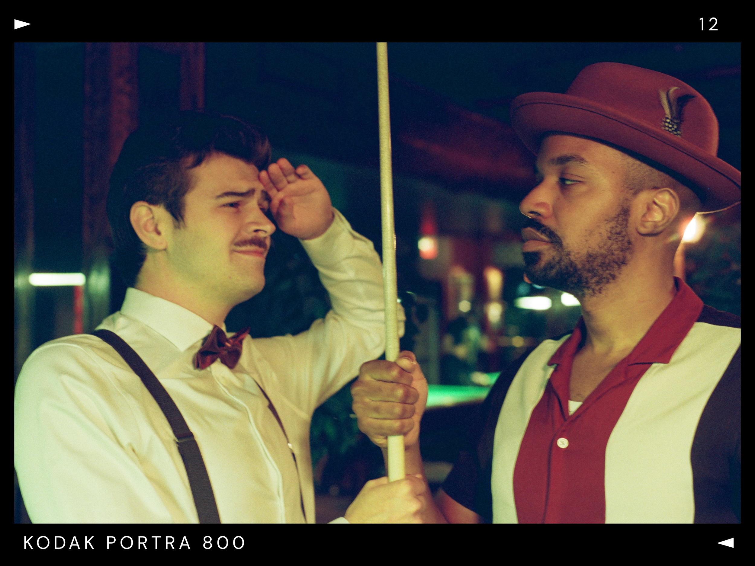 Two men facing each other, one dressed as a bartender with a red hat and shirt, and the other in a white shirt with suspenders and a bow tie, holding a pool cue between them.