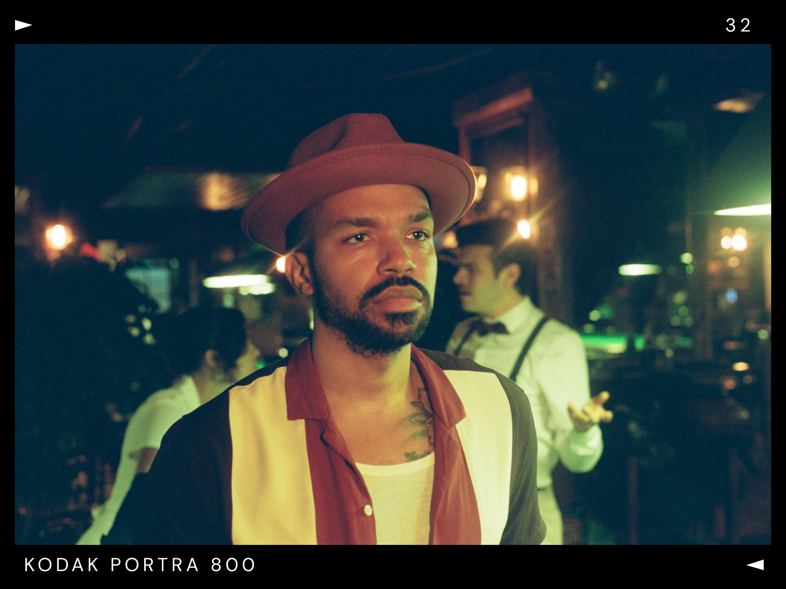 A man with a beard wearing a brown hat, a multicolored jacket, and a white shirt stands in a dimly lit bar or nightclub, with two blurred bartenders or waitstaff in the background.