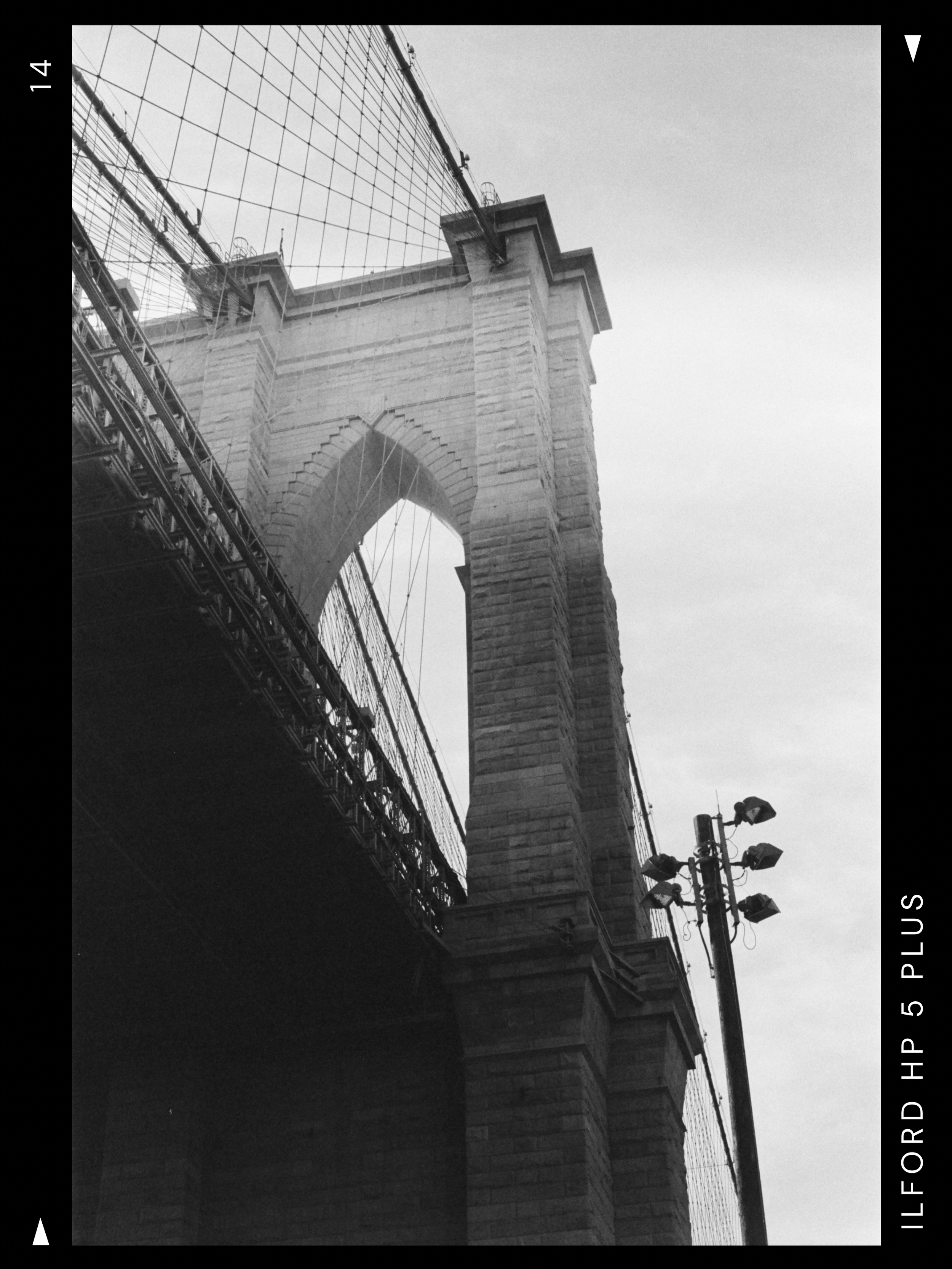 A black and white photograph of the Brooklyn Bridge in New York City, showing its stone tower and suspension cables from a low angle.