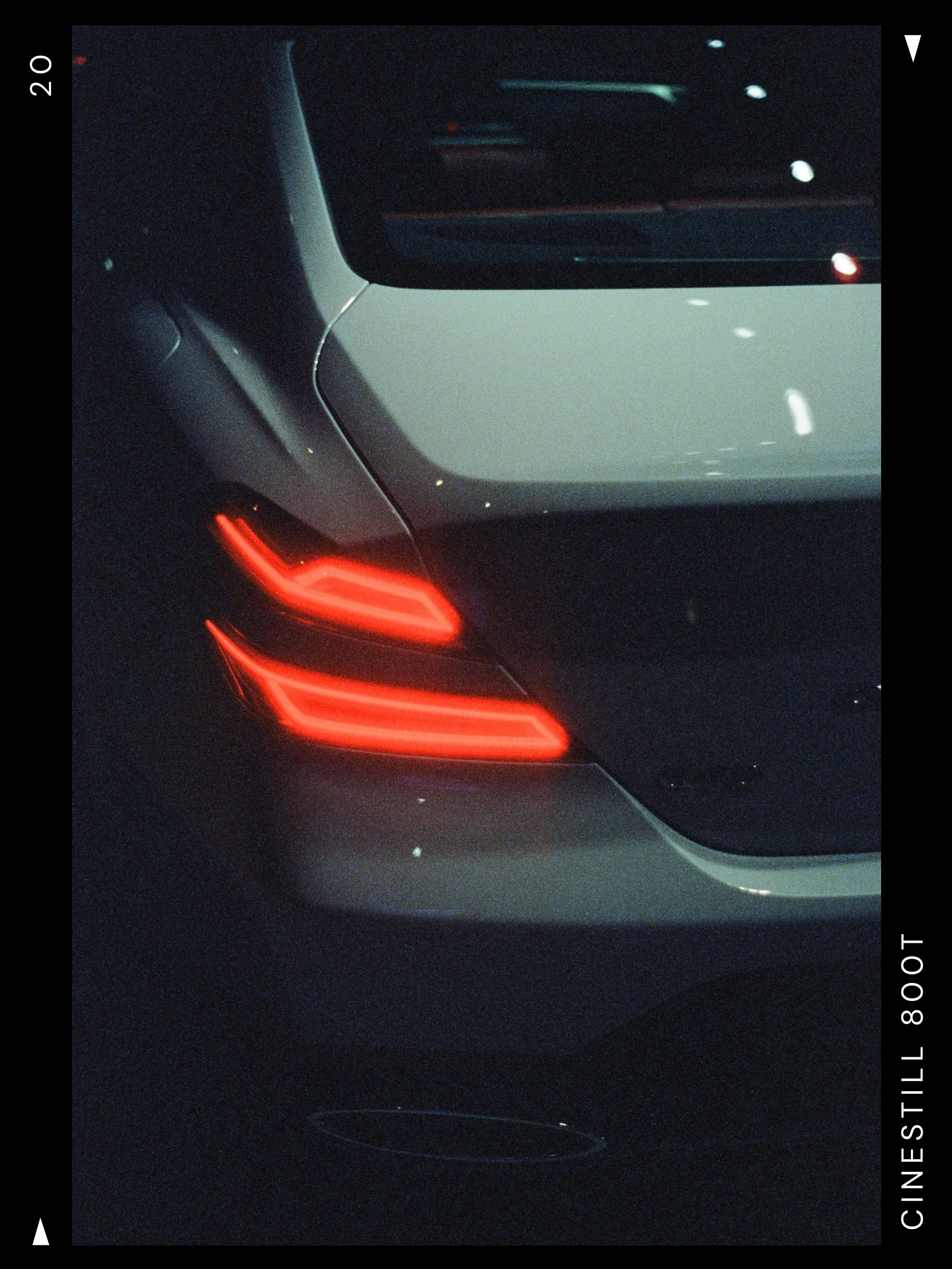 Close-up of the rear corner of a luxury car at night, showing the glowing red tail Lights and part of the white body, with reflections on the car's surface.