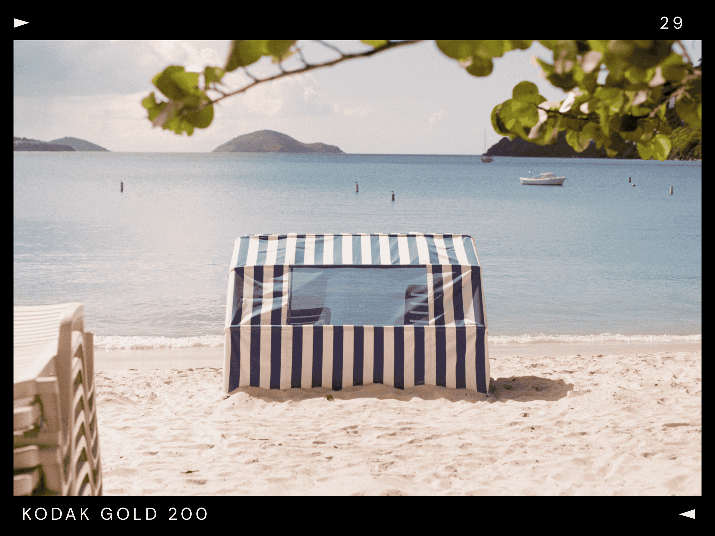 A striped beach tent on sandy shore near calm blue water with small islands and boats in the distance, framed by overhanging tree branches.