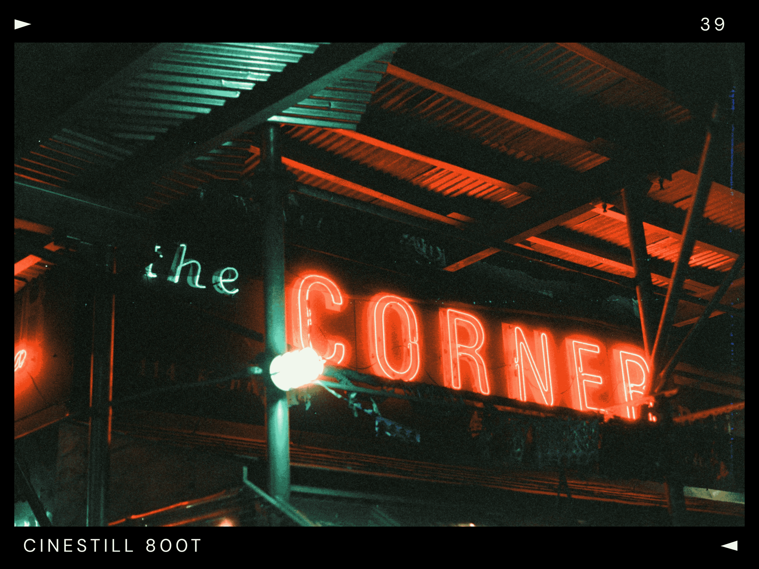 Neon sign reading 'the CORNER' in red and green at night, with metal roofing above and supporting beams.