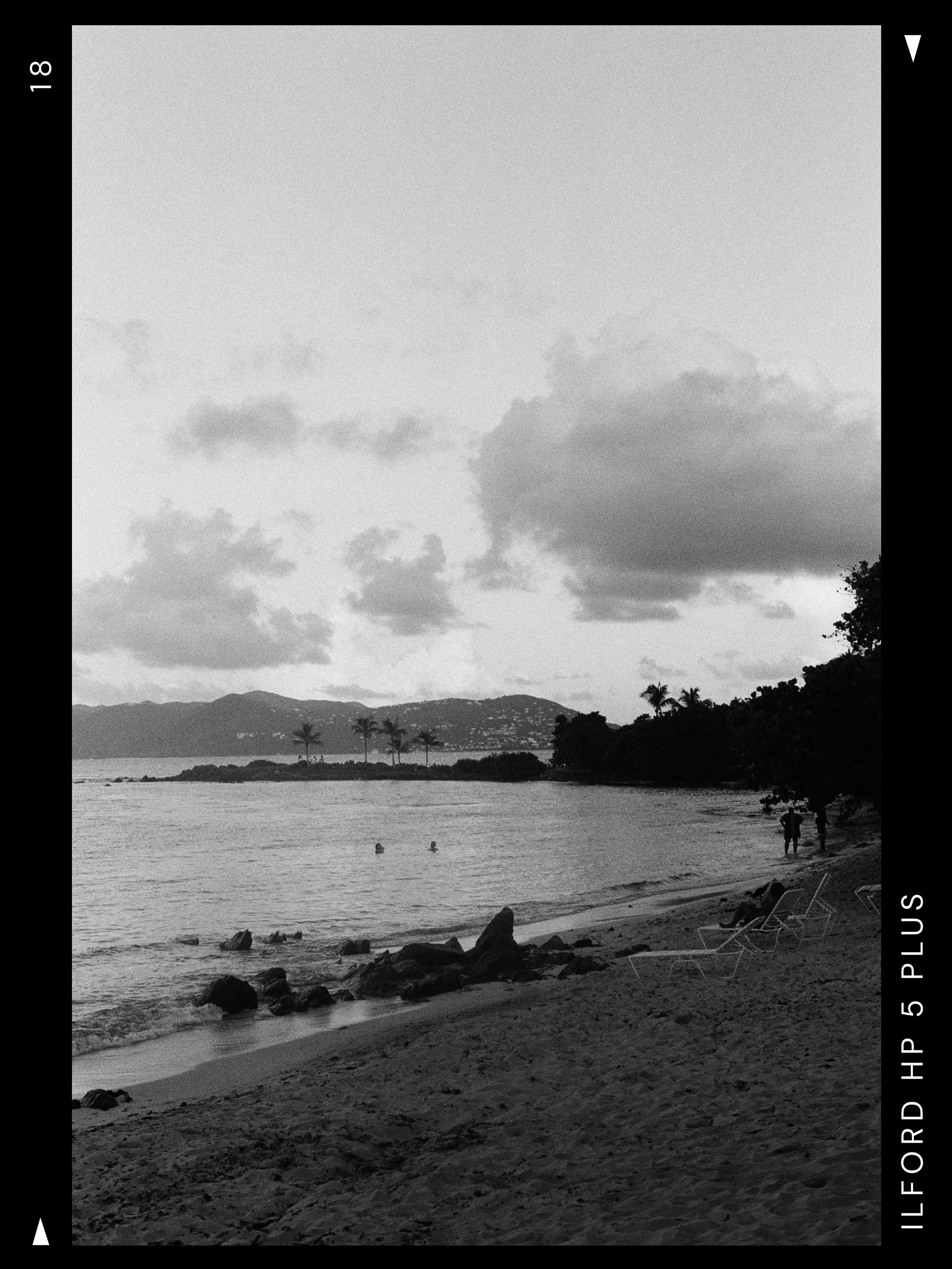 A black and white photo of a St. Thomas beach with rocks and lounge chairs on the sand, a few people swimming in the water, and two people walking along the shoreline, with palm trees, mountains and a cloudy sky in the background.