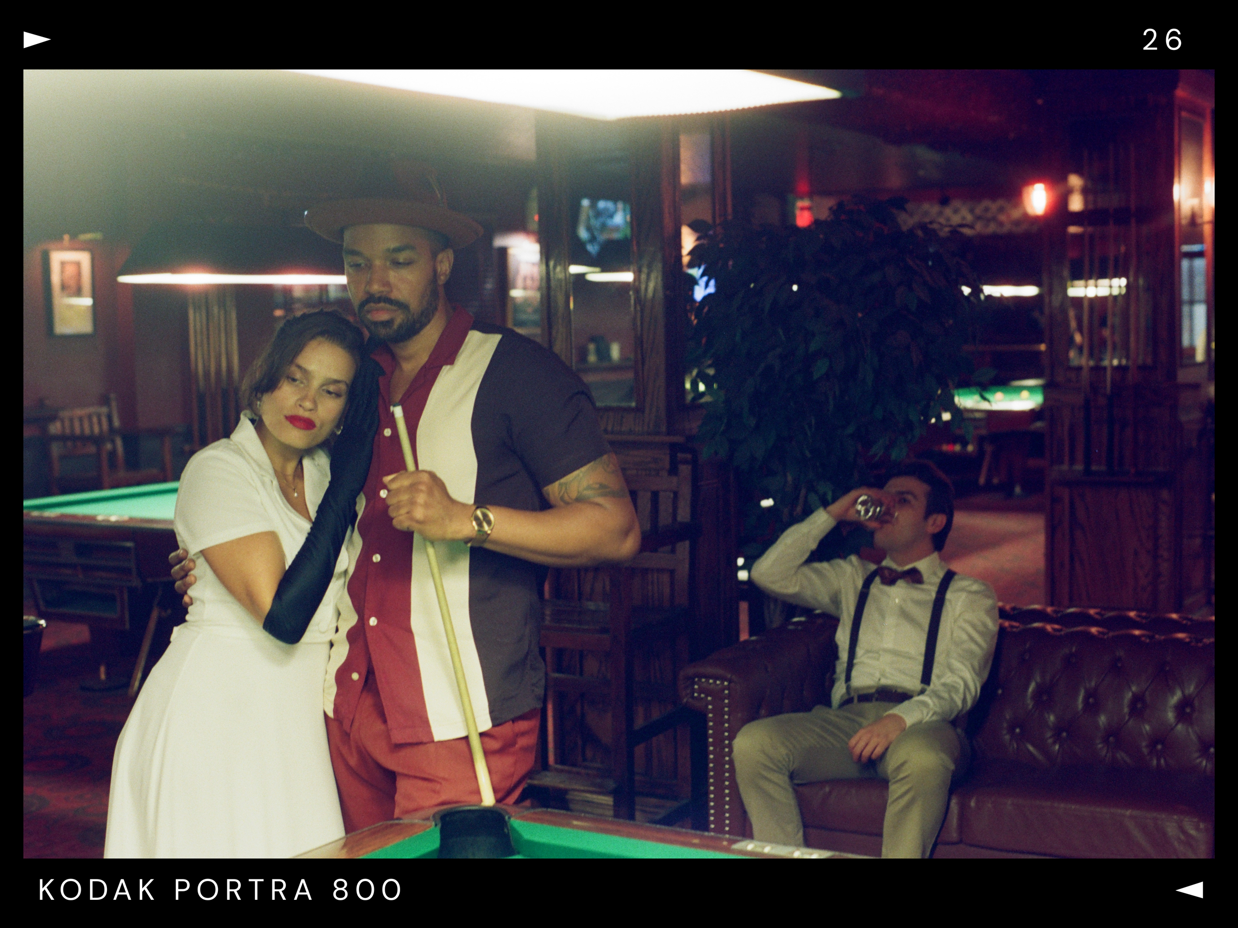 A woman in a white dress leaning on a man with a pool cue in a dimly lit bar, while a man in suspenders and a bowtie drinks from a bottle on a leather couch in the background.