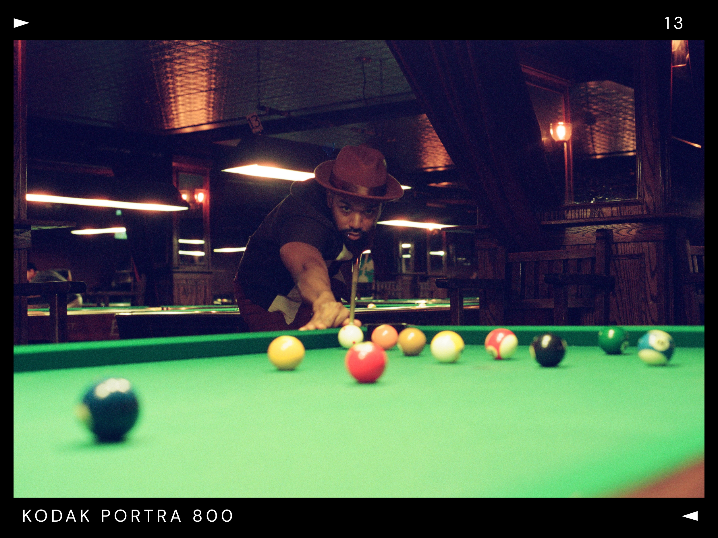 A man in a maroon hat playing pool at a dimly lit billiards hall, leaning over the green table with billiard balls scattered across it.