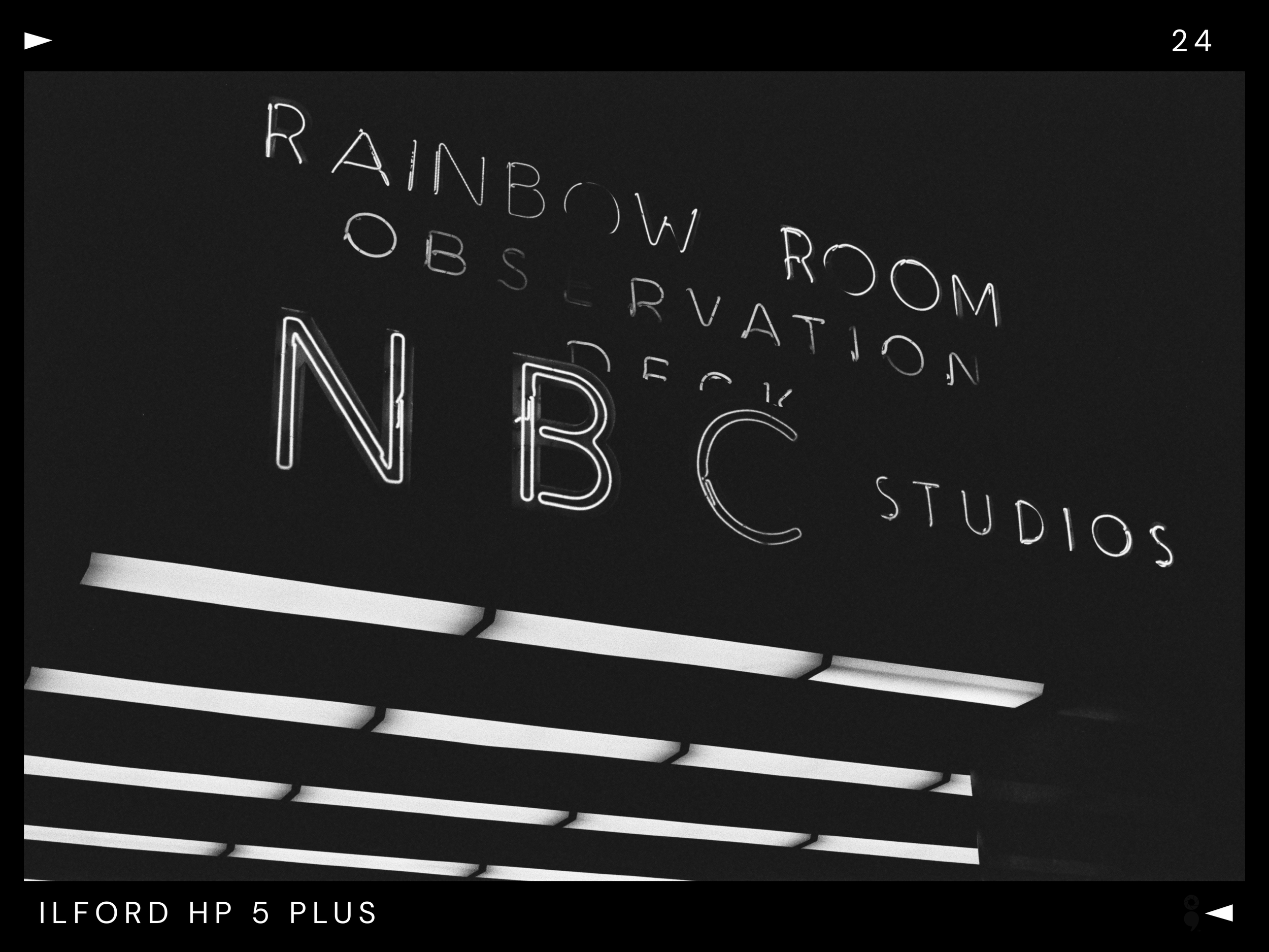 Neon sign reading 'N B C' with words 'Rainbow Room', 'Obstruction', 'Walk', and 'Studios' on a dark background with ceiling lights.