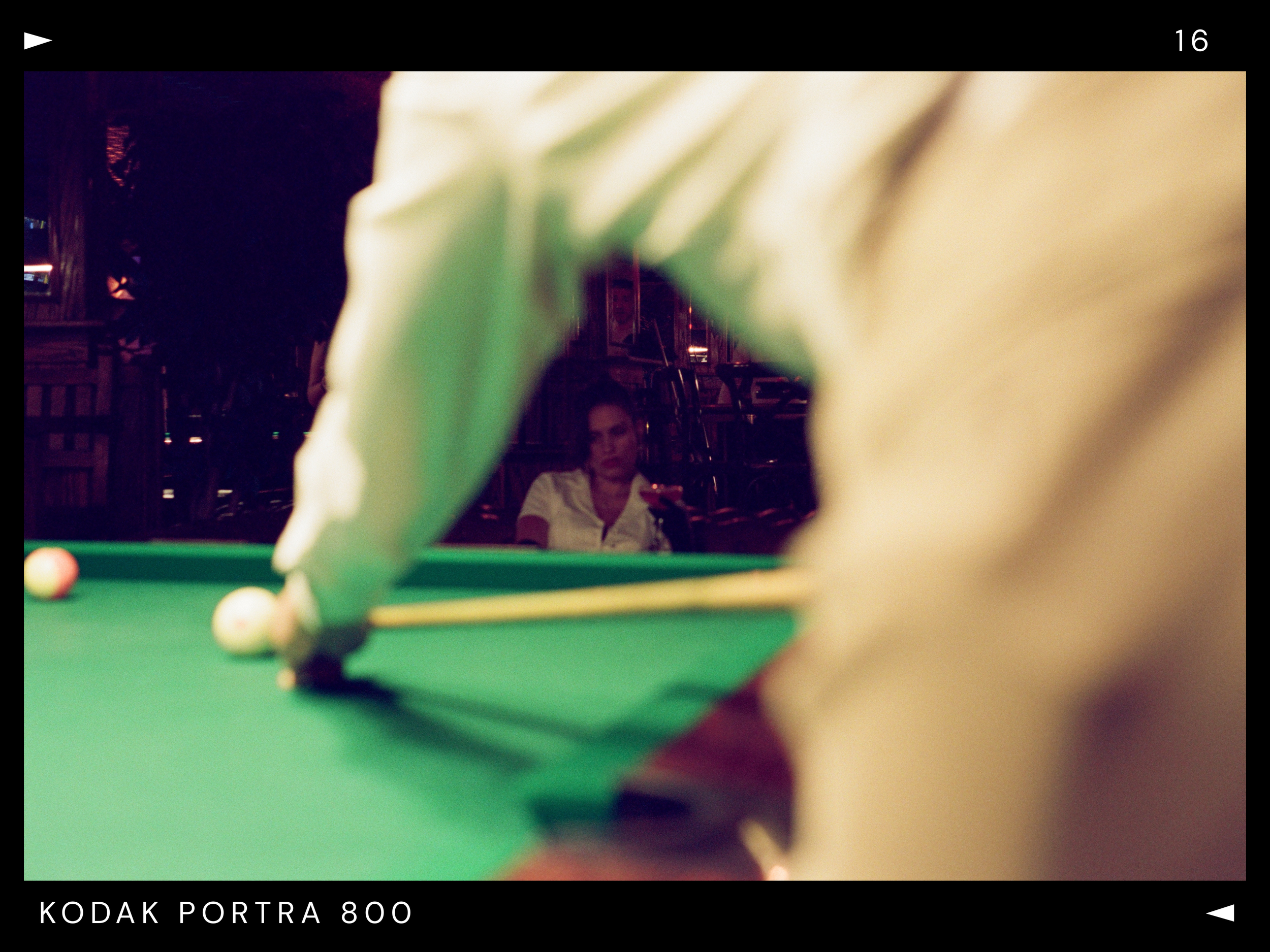 Photo of a person sitting at a pool table, viewed through a blurred foreground, with a woman in a white shirt in the background in a dimly lit room.