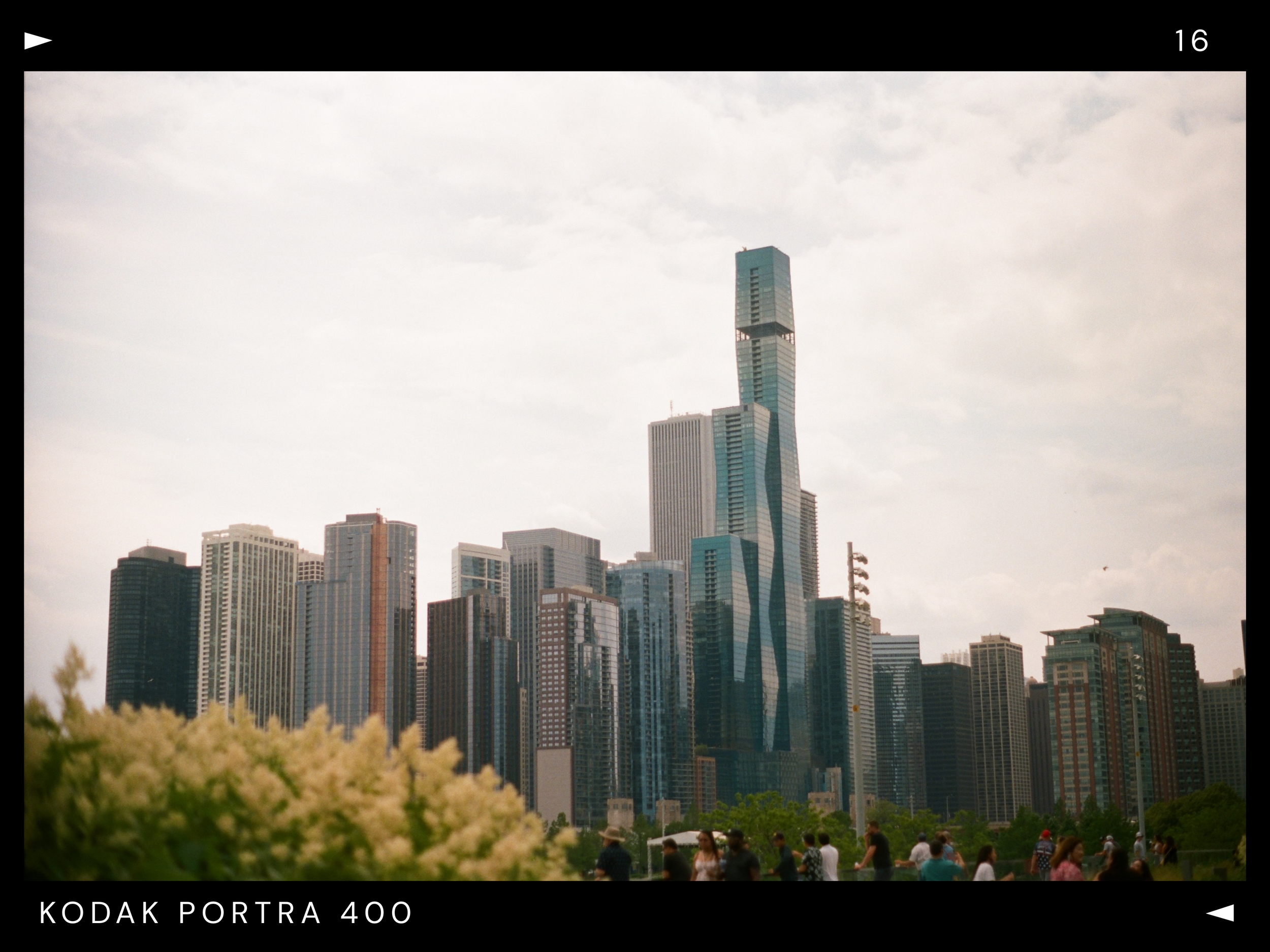 City skyline with tall modern skyscrapers behind a park with trees and people walking on the grass, overcast sky.