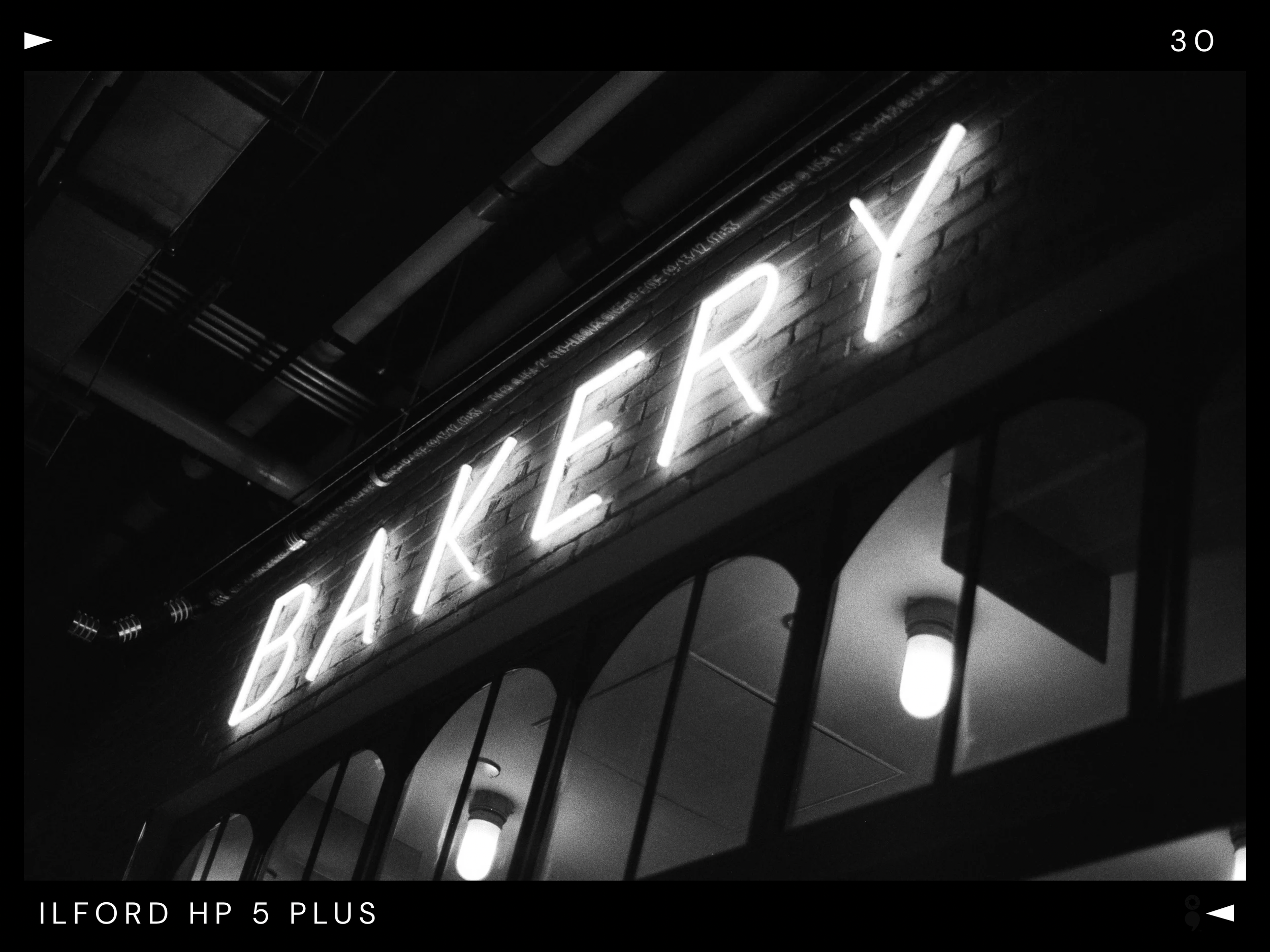 Black and white photo of a neon sign spelling 'BAKERY' on a brick wall, with reflections of ceiling lights and black metal bars in the foreground.