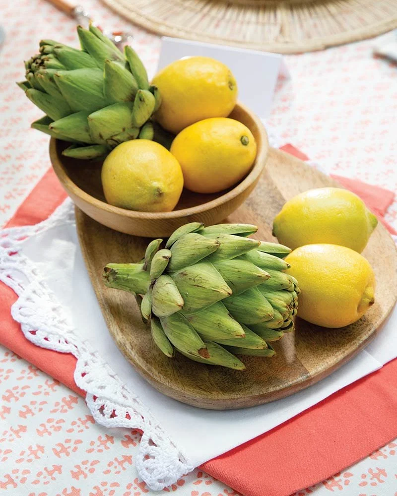 A wooden bowl and a wooden platter each holding green and yellow lemons and a bunch of green artichokes on a table with lace and fabric tablecloths.
