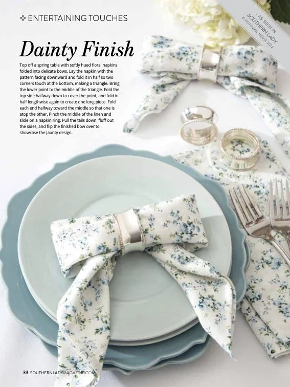 Table setting with pastel blue and white floral dinnerware, a fabric napkin with a floral pattern tied with a silver ring, and champagne flutes and jewelry on a floral tablecloth.