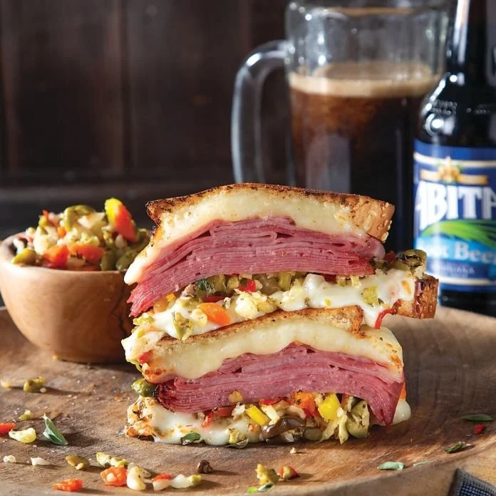 Close-up of a sandwich with ham, cheese, and vegetables on toasted bread, alongside a small bowl of vegetable salad, a pitcher of beer, and a bottle of beer in the background.