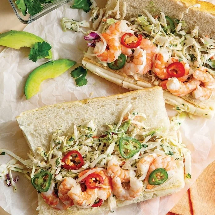 Two shrimp po'boy sandwiches with shredded cabbage, sliced chili peppers, and slices of avocado on the side.