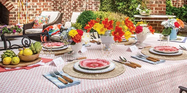 Outdoor patio table set for a meal, decorated with colorful flowers, plates, cutlery, and a pink patterned tablecloth.