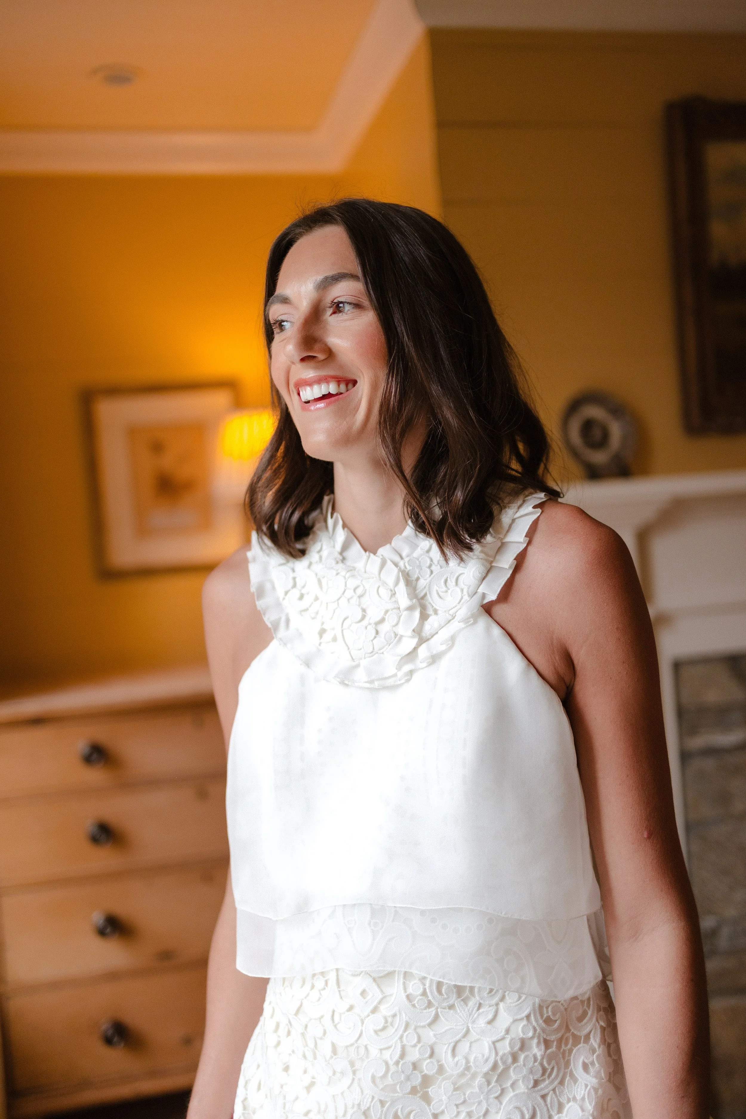 A woman with shoulder-length dark hair, smiling, wearing a white sleeveless dress with ruffled neckline and lace pattern, standing in a warmly lit room with yellow walls, framed artwork, a wooden dresser, and a shelf in the background.