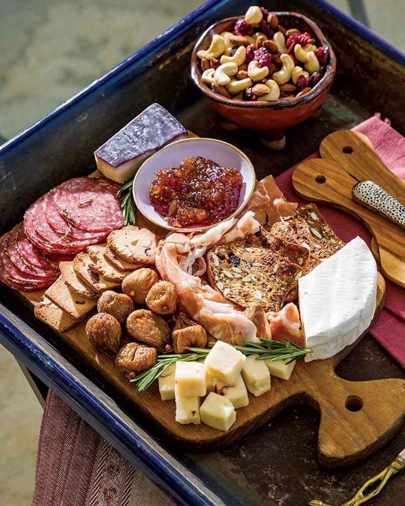 A charcuterie board with various cheeses, cured meats, nuts, dried fruits, fig jam, and breadsticks on a wooden platter.
