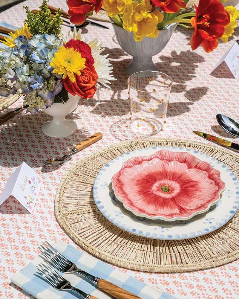 Table setting with floral-themed plates, a bamboo charger, silverware wrapped in a checkered napkin, decorated with floral arrangements in vases, and a pink and white patterned tablecloth.