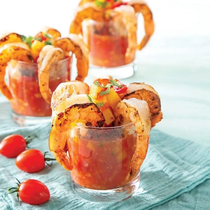 Three glasses of shrimp cocktail with tomato sauce, garnished with lime wedges and chopped vegetables, on a teal tablecloth with cherry tomatoes nearby.