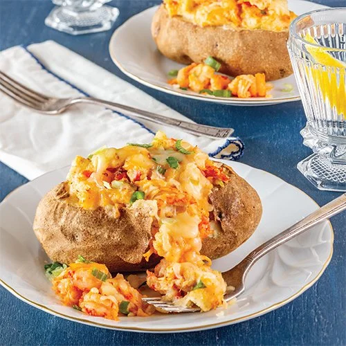 Baked potato topped with melted cheese, bacon bits, and chopped green onions on a white plate with a fork, with another baked potato and a glass of water with lemon in the background.