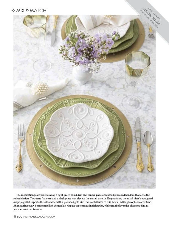 Table setting with green and gold dinnerware, a white embossed plate, gold flatware, a white napkin with a pearl napkin ring, a small floral centerpiece with purple flowers, two wine glasses, and two gold geometric candle holders.