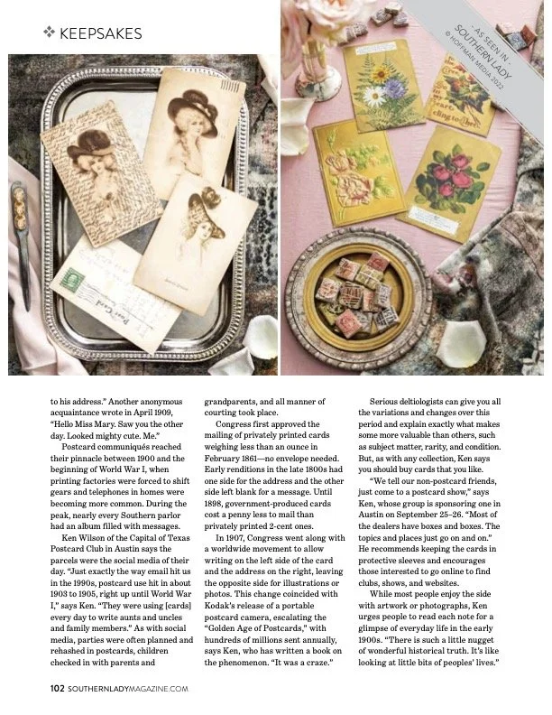 A collection of vintage postcards and greeting cards displayed on trays and a table, with flowers and decorative items around, showcasing historical and artistic designs.