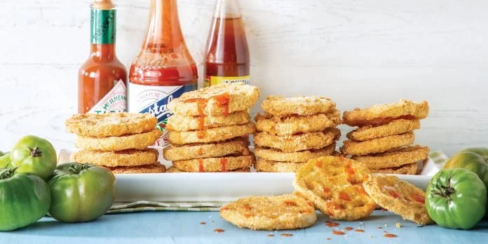 Stacked fried chicken fillets with ketchup bottles and green tomatoes on a white platter.
