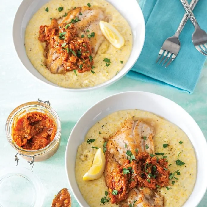 Two bowls of fish fillets served on creamy polenta with tomato-based sauce and lemon wedges, alongside a small jar of additional sauce.