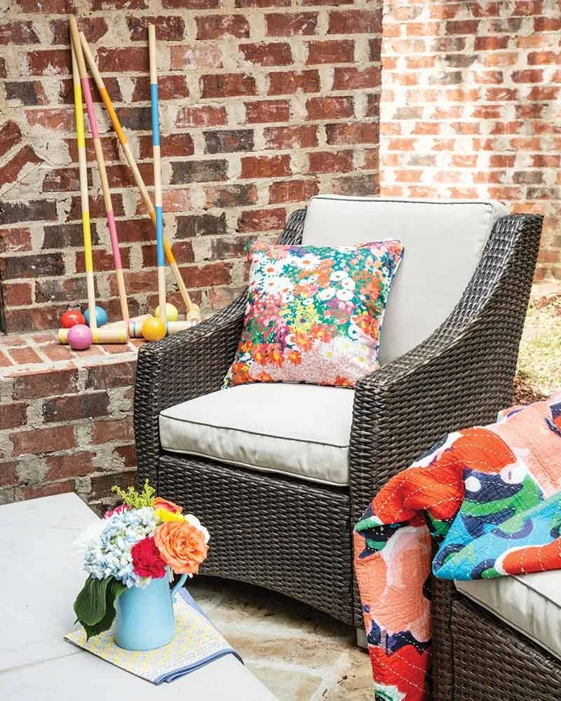 A wicker outdoor chair with white cushions, a colorful floral pillow, next to a small table with a flower arrangement, and a brick wall in the background with colorful croquet mallets leaning against it.