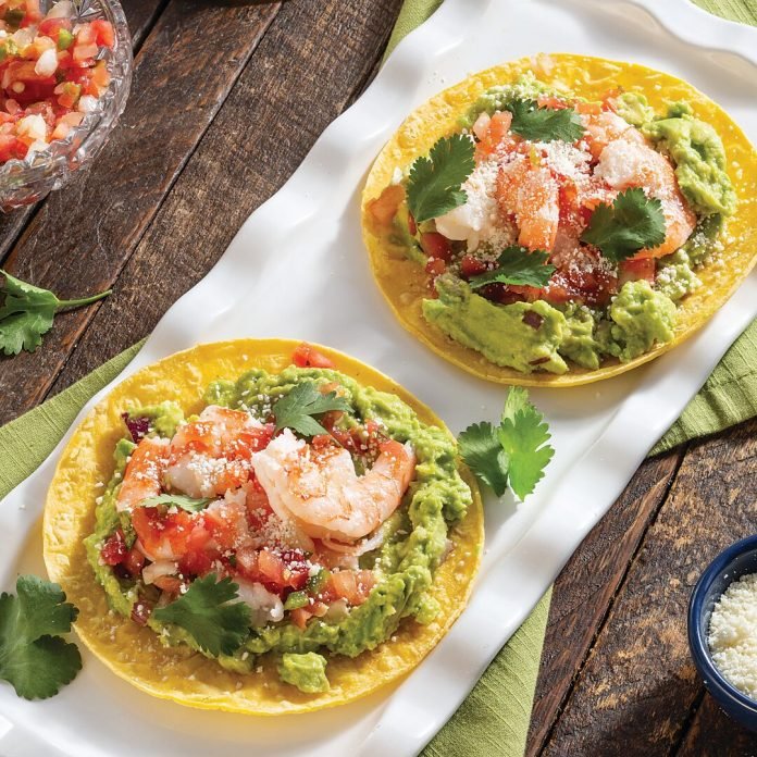 Two fish tacos with shrimp, guacamole, diced tomatoes, shredded cheese, and cilantro on yellow corn tortillas, served on a white platter with a rustic wooden table background.