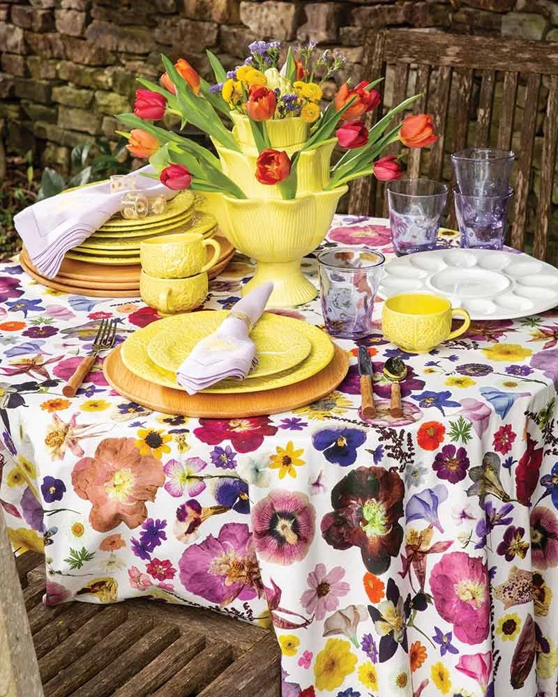 A table set for outdoor dining with a floral tablecloth, yellow dishware, a large yellow vase with tulips and other flowers, and glassware, against a stone wall background.