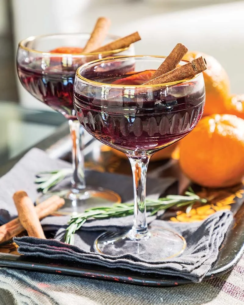Two glasses of red wine garnished with cinnamon sticks on a tray, with oranges and sprigs of rosemary nearby.