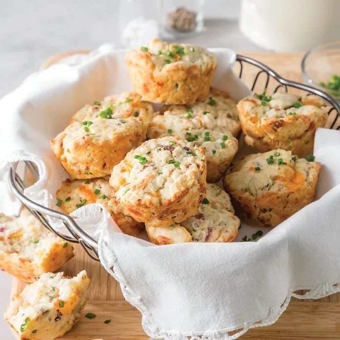 A wire basket filled with savory breakfast muffins garnished with chopped chives, sitting on a white cloth-lined wooden board.