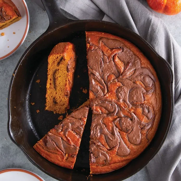 A round apple cake with a slice cut out, topped with chocolate glaze, in a black baking pan on a gray surface with a gray cloth nearby.