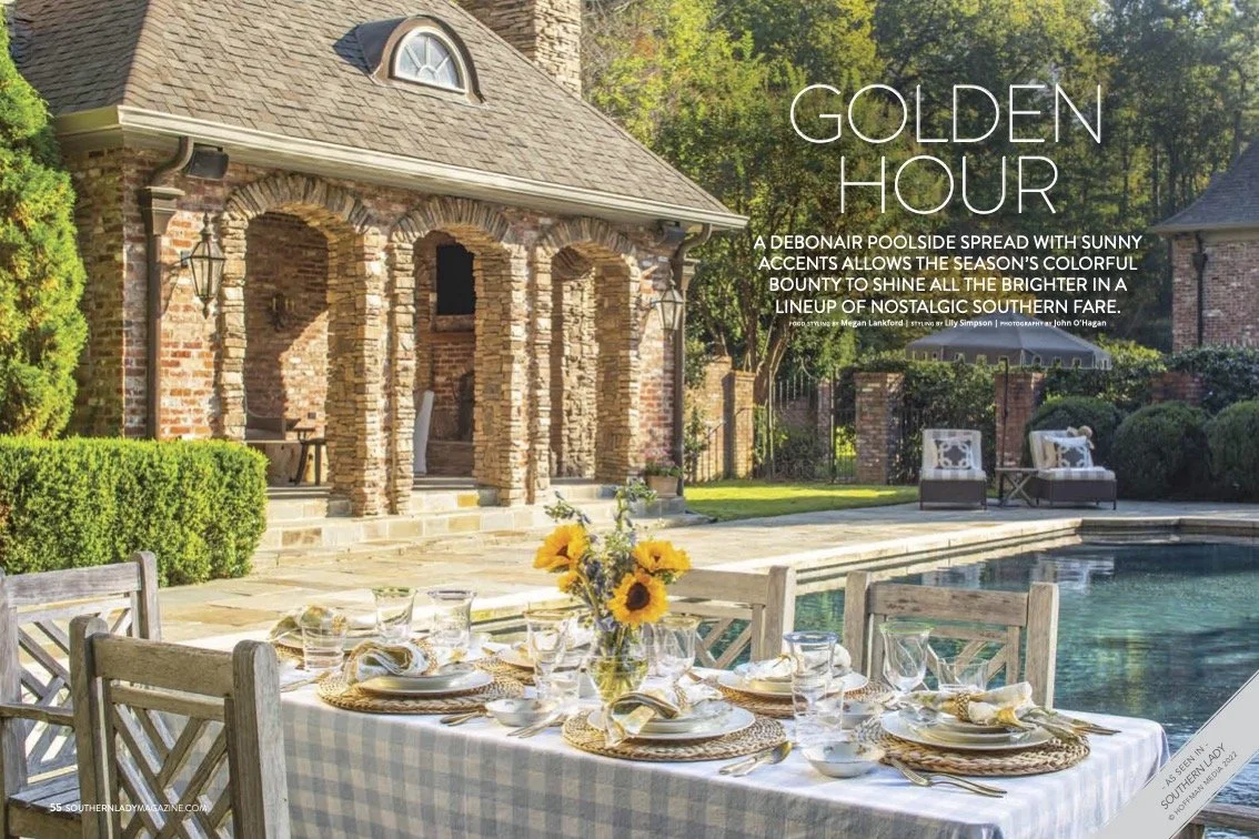 Outdoor dining table set by a swimming pool, with sunflowers centerpiece and place settings, in front of a brick house with arched brick porch and lush greenery.