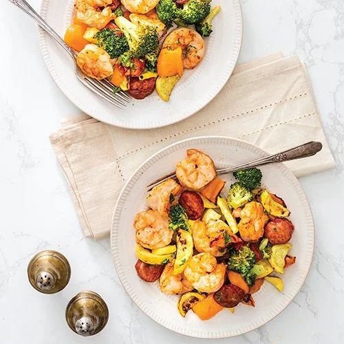 Two plates of shrimp and vegetable stir-fry with broccoli, yellow squash, and cherry tomatoes, on a white marble surface with salt and pepper shakers nearby.